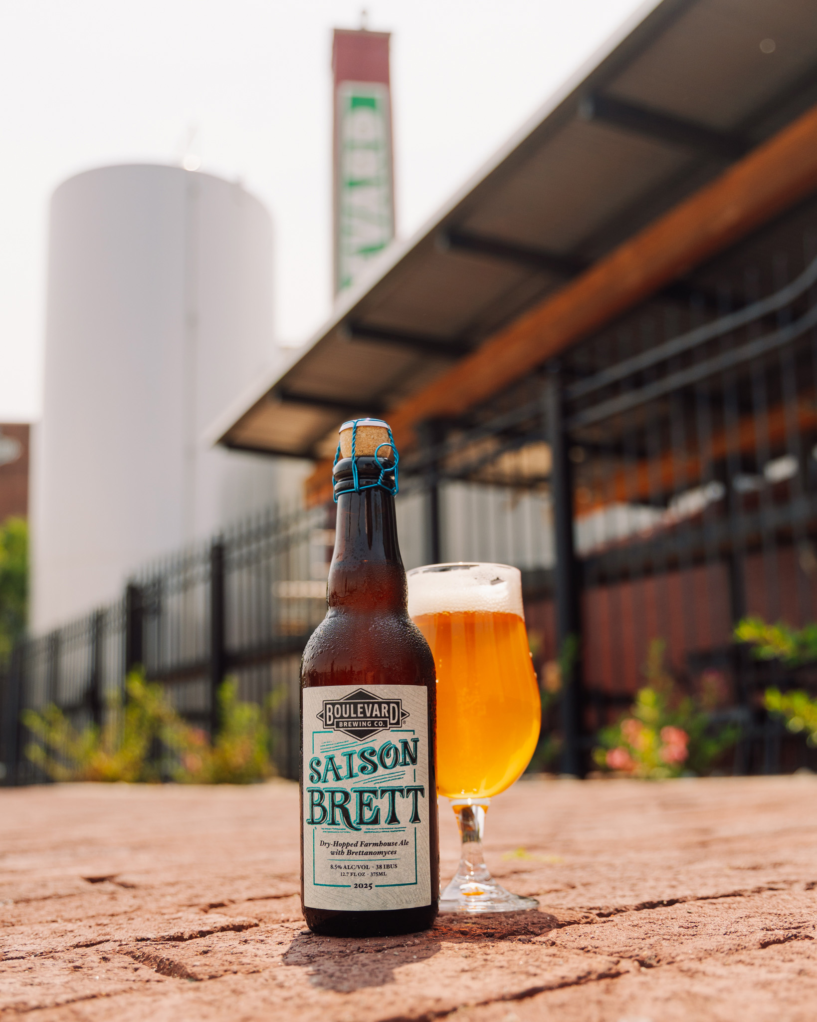 A bottle of Boulevard Saison Brett beer and a filled glass are set on a stone surface outdoors, with a brewery building and large fermenter tank visible in the background.