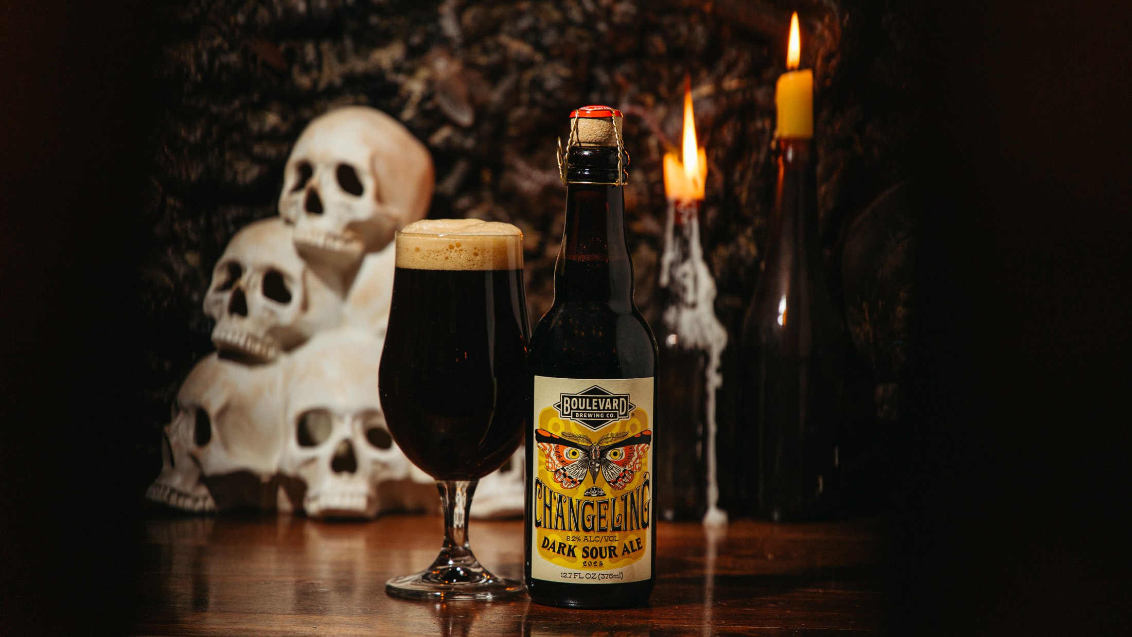 A bottle of Boulevard Changeling Dark Sour Ale and a full glass sit on a table. In the background are lit candles and a stack of decorative skulls, creating a moody, Halloween-like atmosphere.