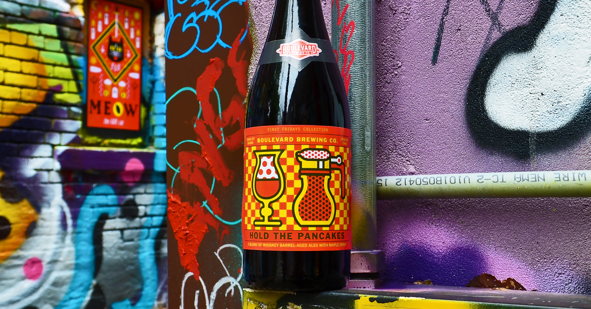 A bottle of Boulevard Brewing Co.'s "Hold the Pancakes" beer stands against a colorful, graffiti-covered wall. The label features vibrant red and yellow patterns with images of a waffle and a beer glass.