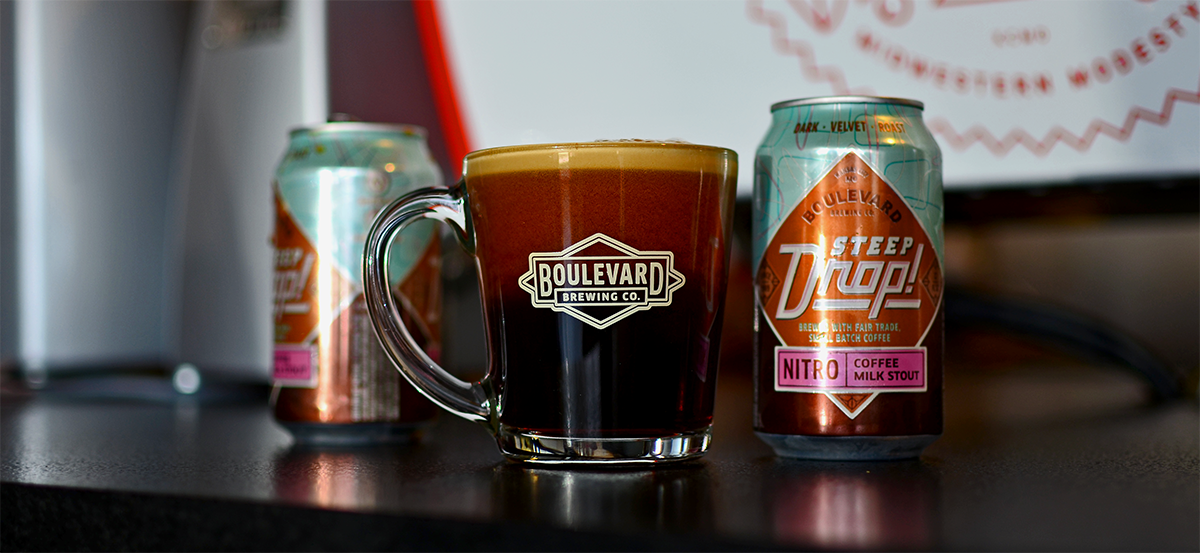 A close-up of a glass mug filled with dark coffee milk stout, featuring the Boulevard Brewing Co. logo. Two cans of Steep Drop Coffee Milk Stout are in the background, one open and one closed, placed on a dark counter.