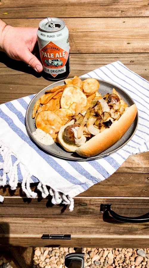 A hand holds a can of pale ale beer next to a plate on a blue and white striped cloth. The plate contains a sausage in a bun topped with grilled onions and a serving of potato chips. This is all set on a wooden table outdoors.