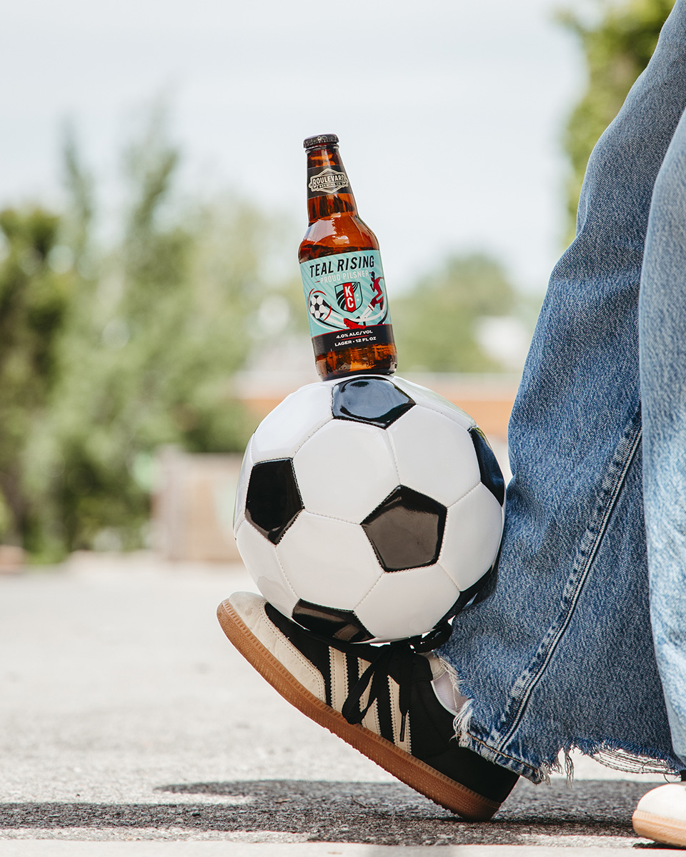 A person wearing jeans and black sneakers balances a soccer ball with their foot. A bottle of beer labeled "Teal Rising" is placed on top of the ball. The background is outdoors and blurry.