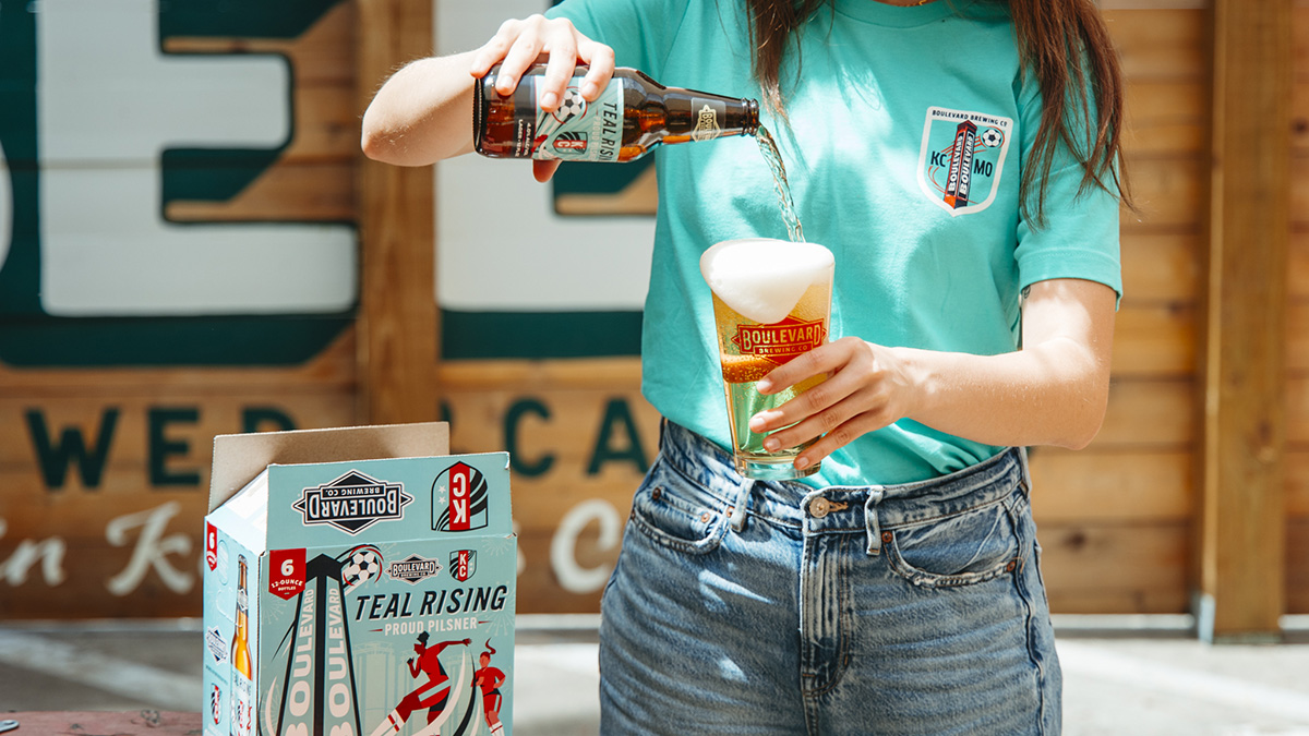 A person in a teal T-shirt pours beer from a bottle into a glass with a large foamy head. An open six-pack of Boulevard Brewing Co. beer sits on a table in front of them.