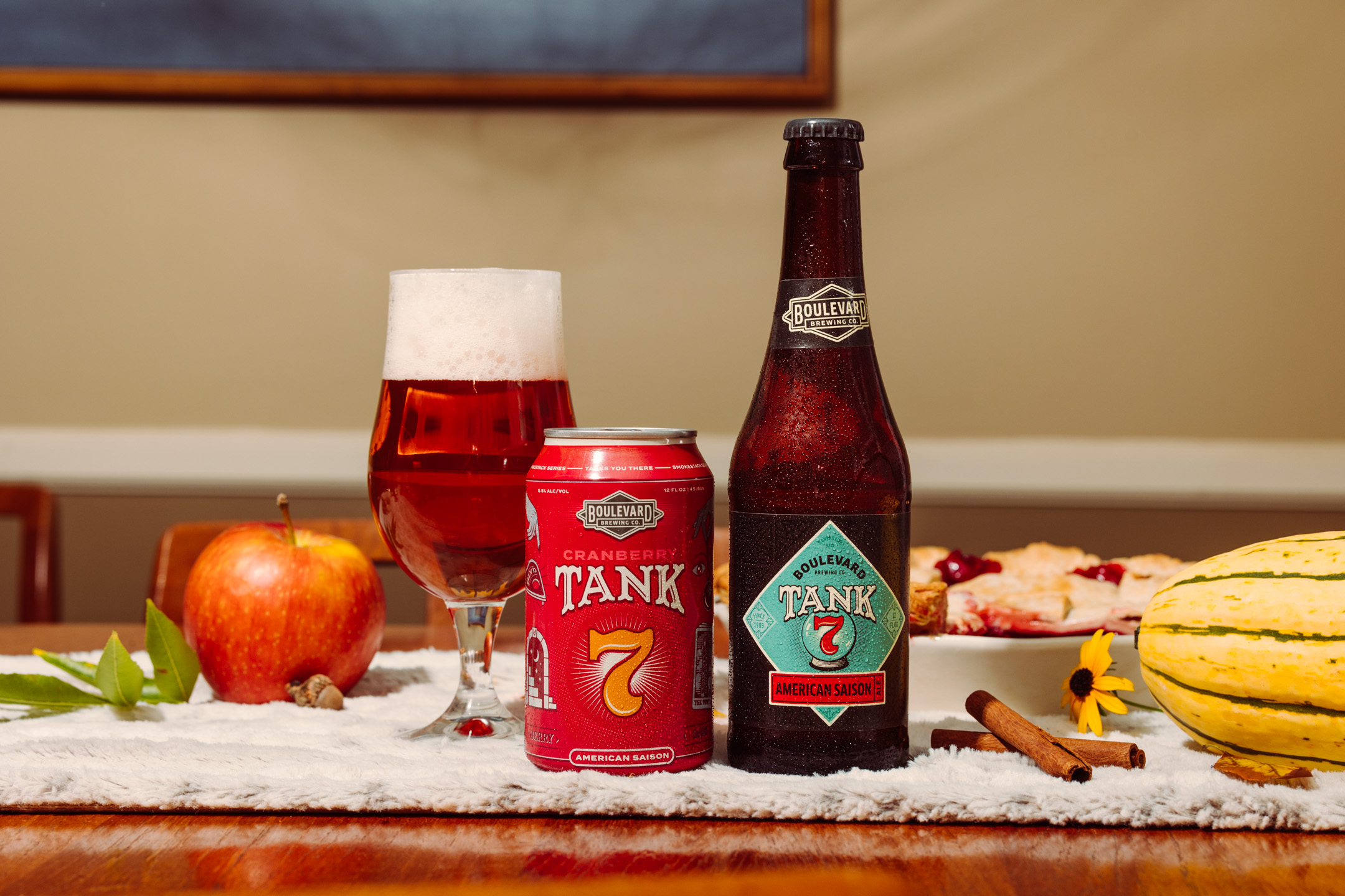 A can and bottle of Boulevard Tank 7 Cranberry Saison beer sit on a table with a filled glass, surrounded by an apple, squash, cinnamon stick, pie, and fall decorations.