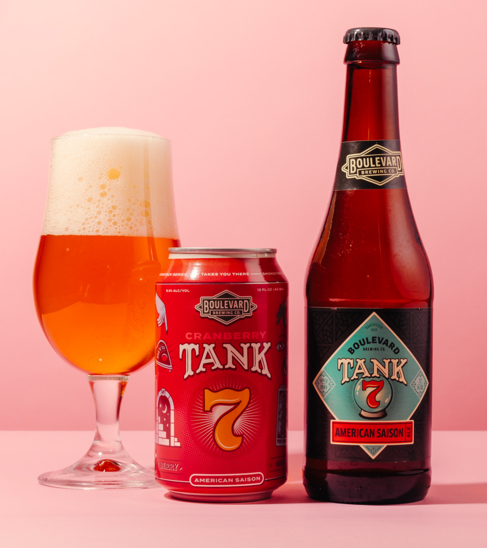 A glass of amber beer with a foamy head stands next to a red can and a brown bottle of Boulevard Brewing Co. Tank 7 American Saison, against a pink background.