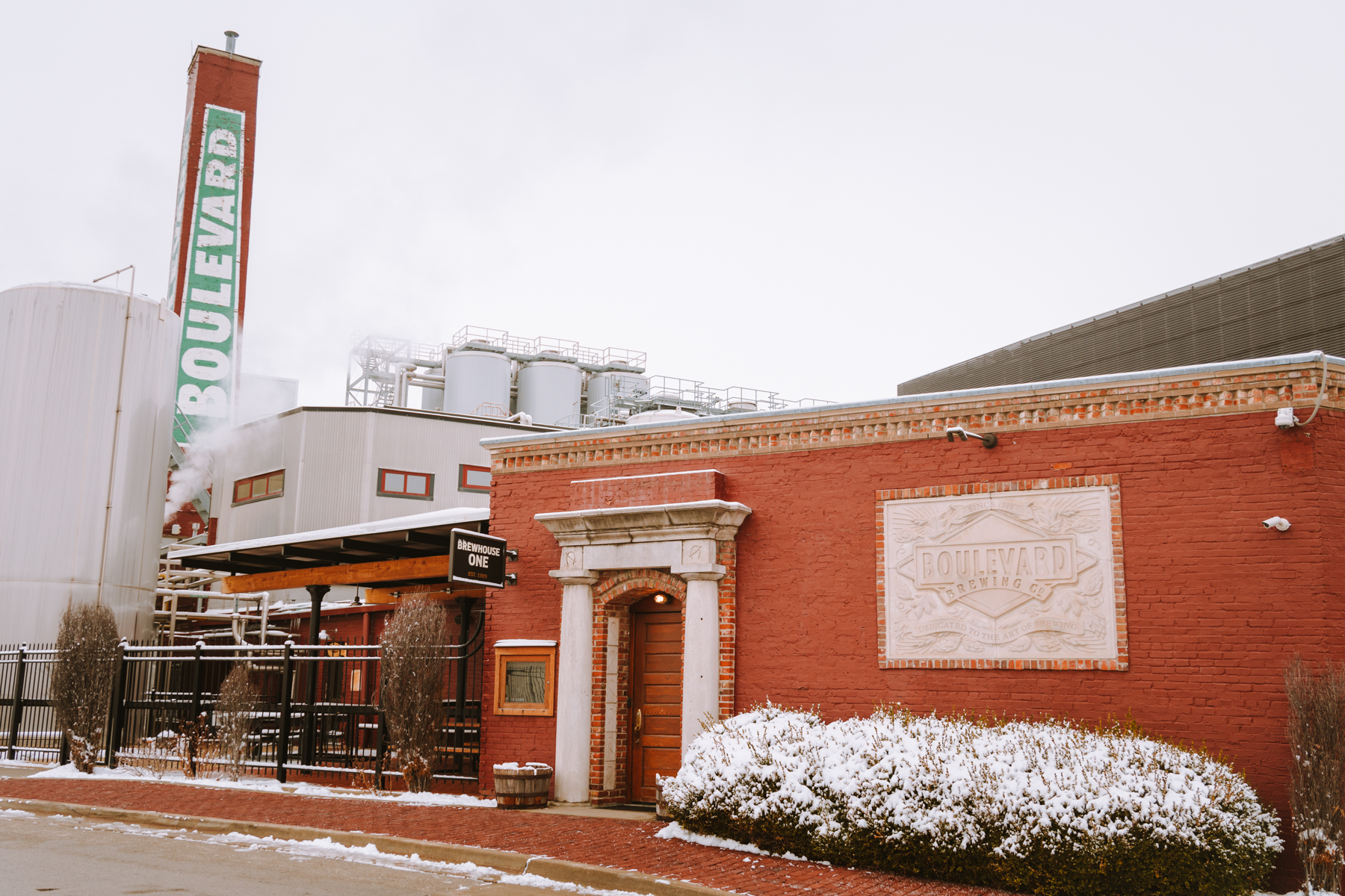 A red brick building with a “Boulevard” sign and engraved plaque stands next to brewing tanks, with light snow on the ground and bushes, under a cloudy sky.