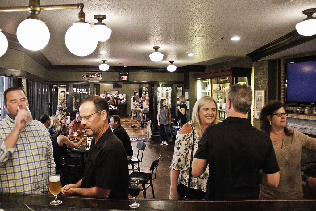 A lively bar scene with people chatting, drinking, and socializing. Groups sit at tables and stand near the bar, under warm lighting and globe-shaped ceiling lights. The atmosphere appears relaxed and friendly.