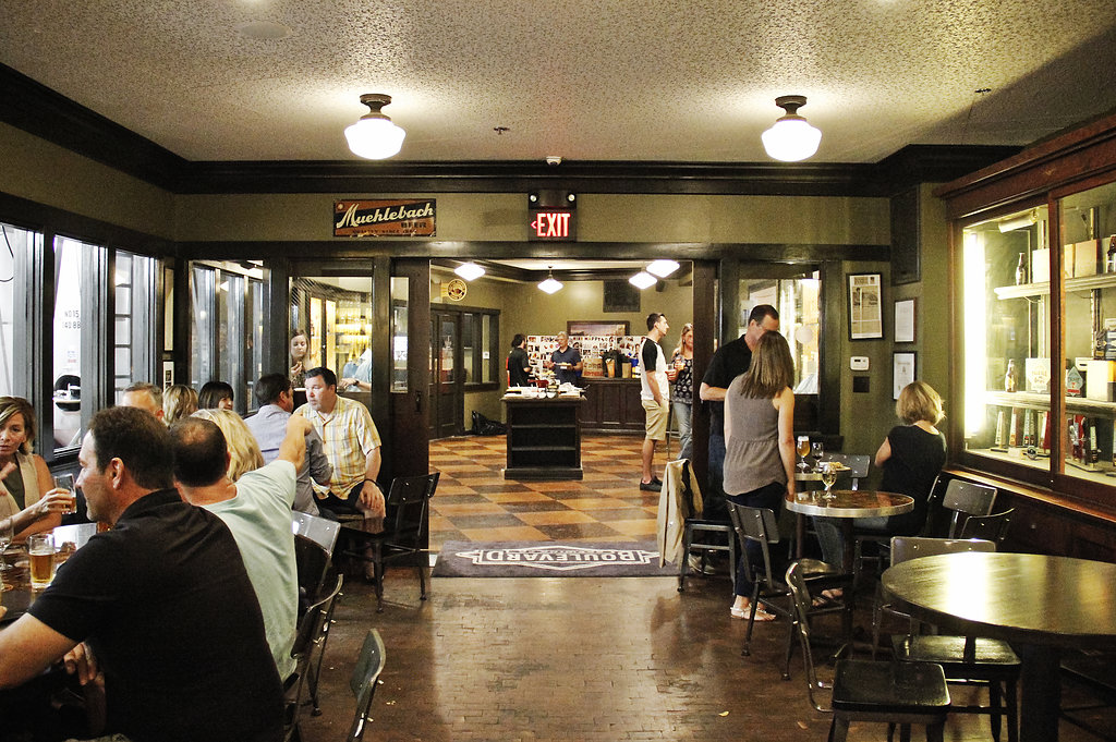 People sit at tables inside a warmly lit bar or restaurant with glass display cases on the right, decorative tile floor, and an open area leading to another section in the background. Some guests are eating, drinking, and talking.
