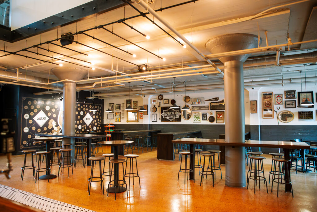 A modern, spacious bar with tall tables and stools, industrial-style lighting, and walls decorated with framed pictures and brewery signs. The floor is polished and orange-toned, and the overall atmosphere is casual and inviting.