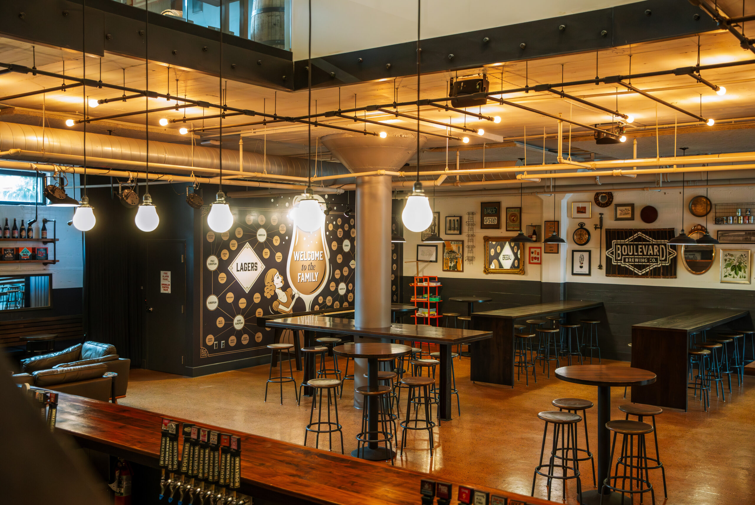 A modern brewery taproom with high tables and stools, exposed pipes, hanging lights, wall art, and signage. The space has a warm, industrial feel with wooden accents and various beer-related decorations.