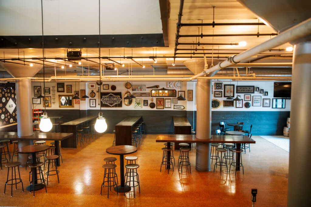 A spacious, modern bar with high tables and stools. Warm lighting hangs from the ceiling. Art and signs cover the back wall. The bar has an industrial style with exposed pipes and a polished floor. No people are present.