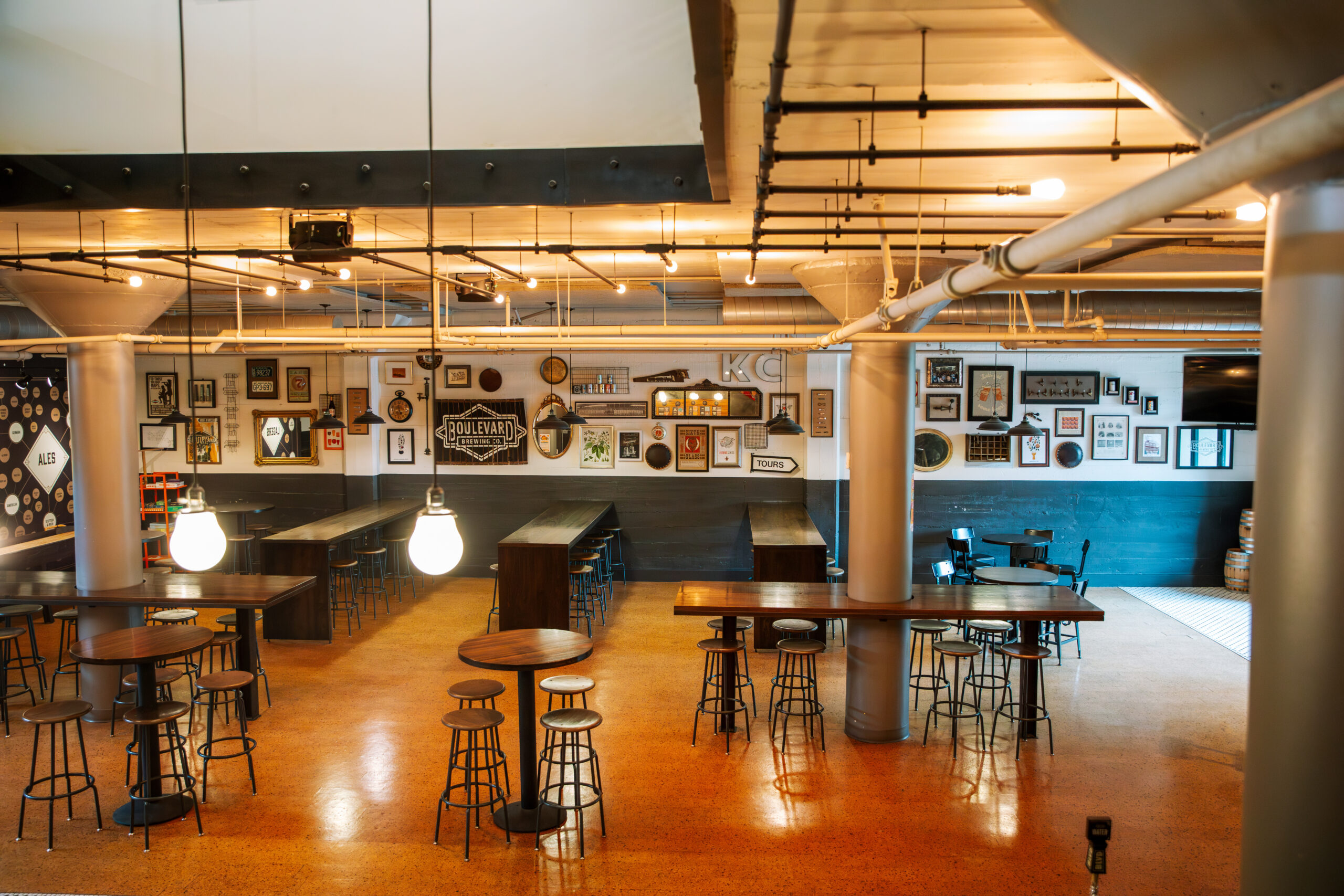 A spacious, modern bar with high tables and stools. Warm lighting hangs from the ceiling. Art and signs cover the back wall. The bar has an industrial style with exposed pipes and a polished floor. No people are present.