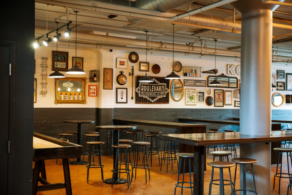 A modern, industrial-style bar with high tables and stools. The walls are decorated with framed pictures, mirrors, and a “Boulevard” sign. Warm lighting creates a cozy atmosphere. The bar appears empty.