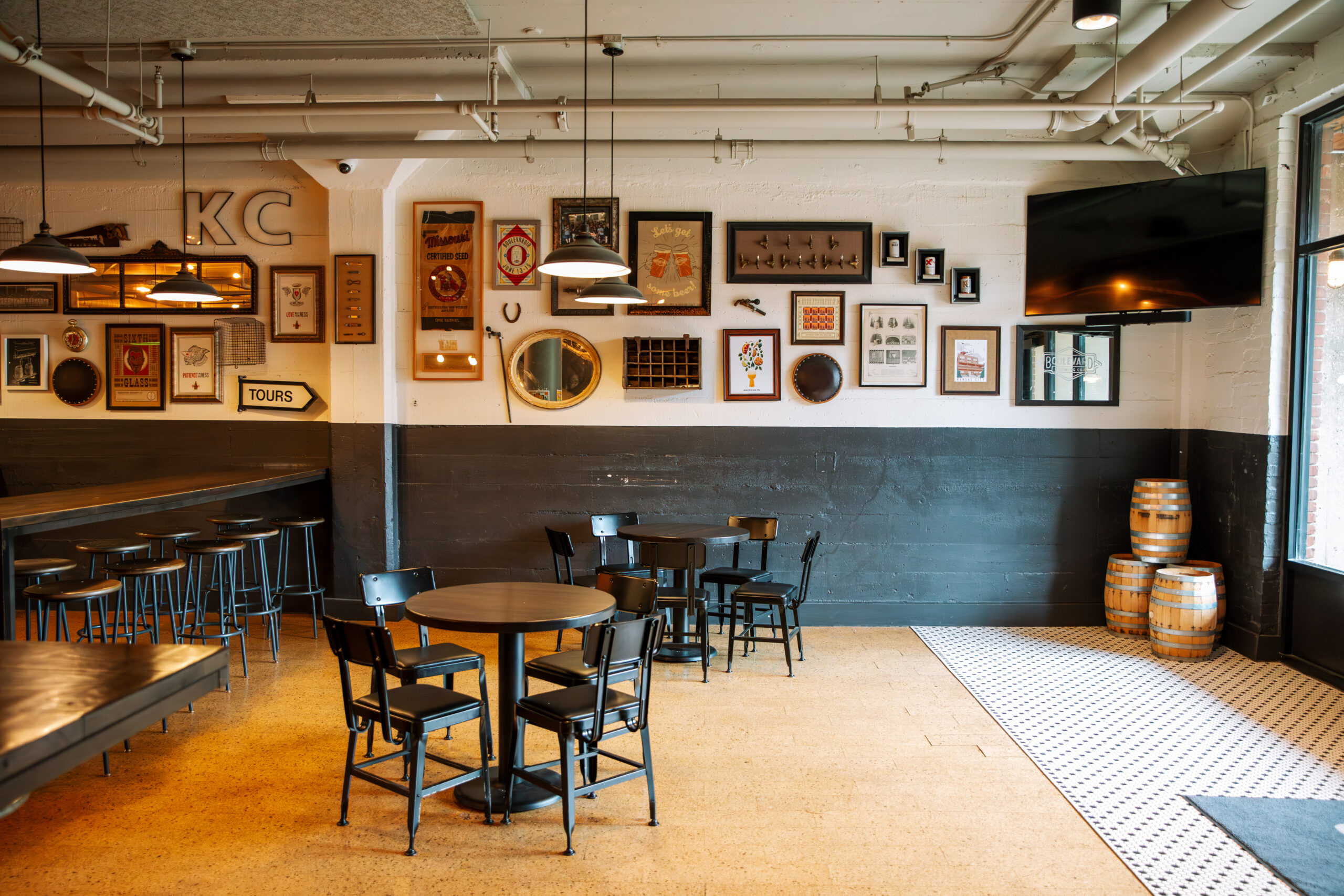 A cozy, modern bar with black tables and chairs, framed art and memorabilia on white walls, hanging lights, stacked barrels, and a large TV in the corner near tall windows. No people are present.