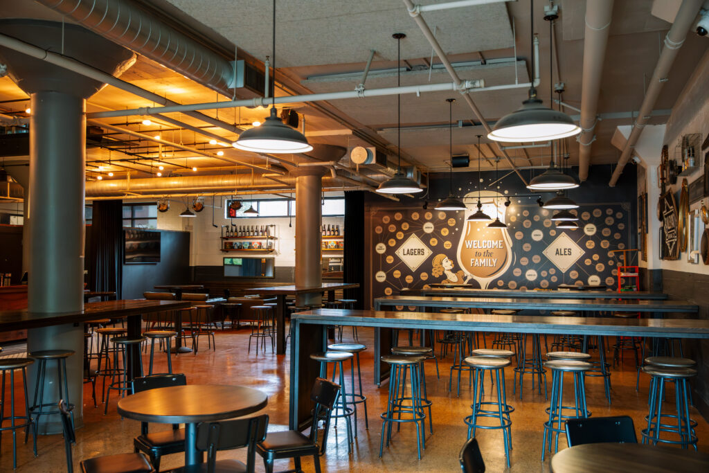 A spacious, modern brewery taproom with high ceilings, exposed pipes, wooden tables, and metal stools. A mural on the wall reads “Welcome to the family” and features beer taps labeled lagers and ales.