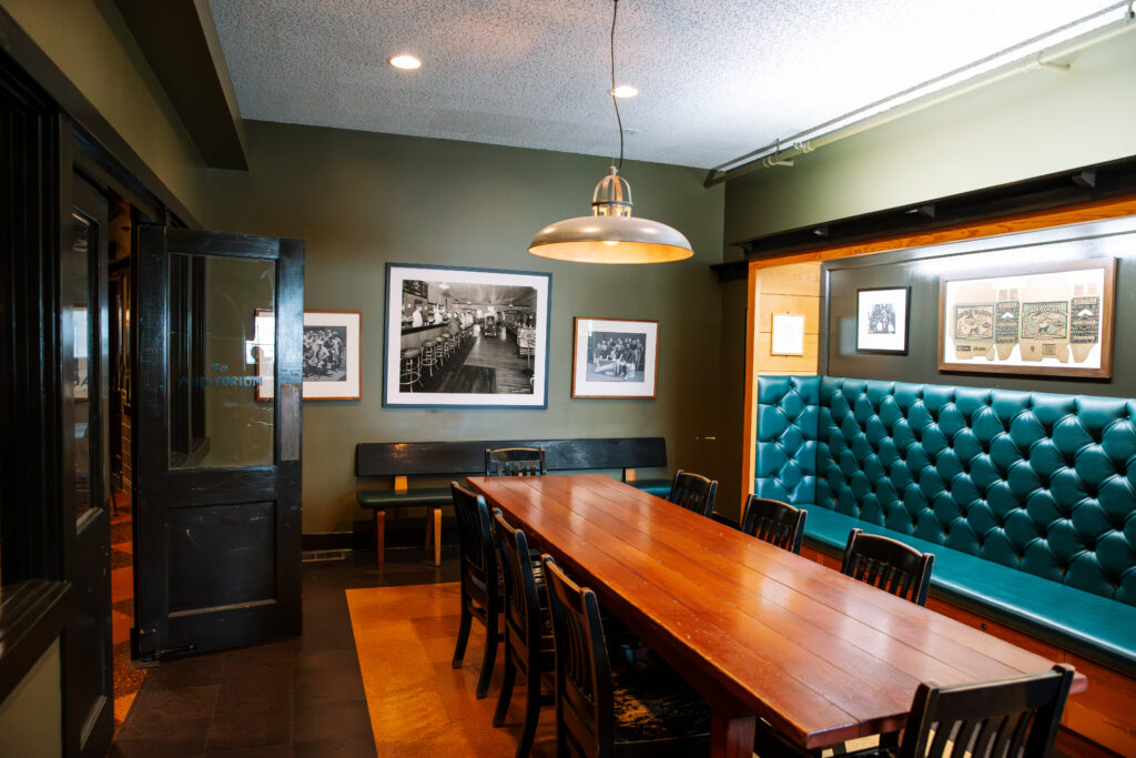 A cozy dining room with a long wooden table surrounded by chairs, a green tufted bench along one wall, framed black-and-white photos, and a hanging light fixture above the table.