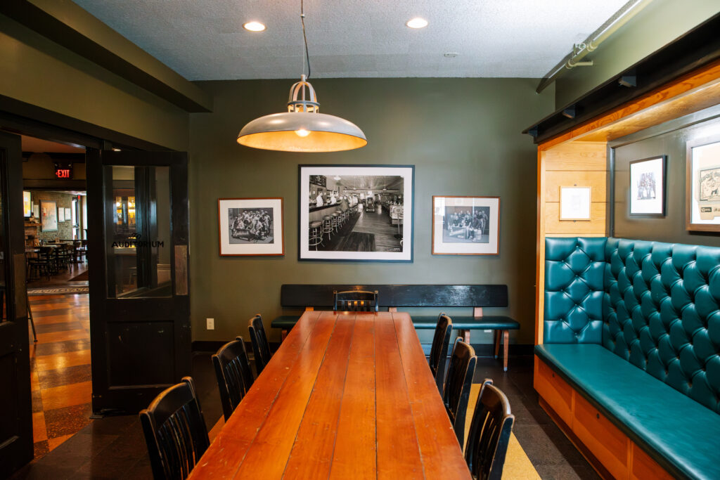 A long wooden dining table with black chairs is centered in a room, with a teal tufted bench along one wall and framed black-and-white photos hanging above it. A large overhead lamp lights the space.