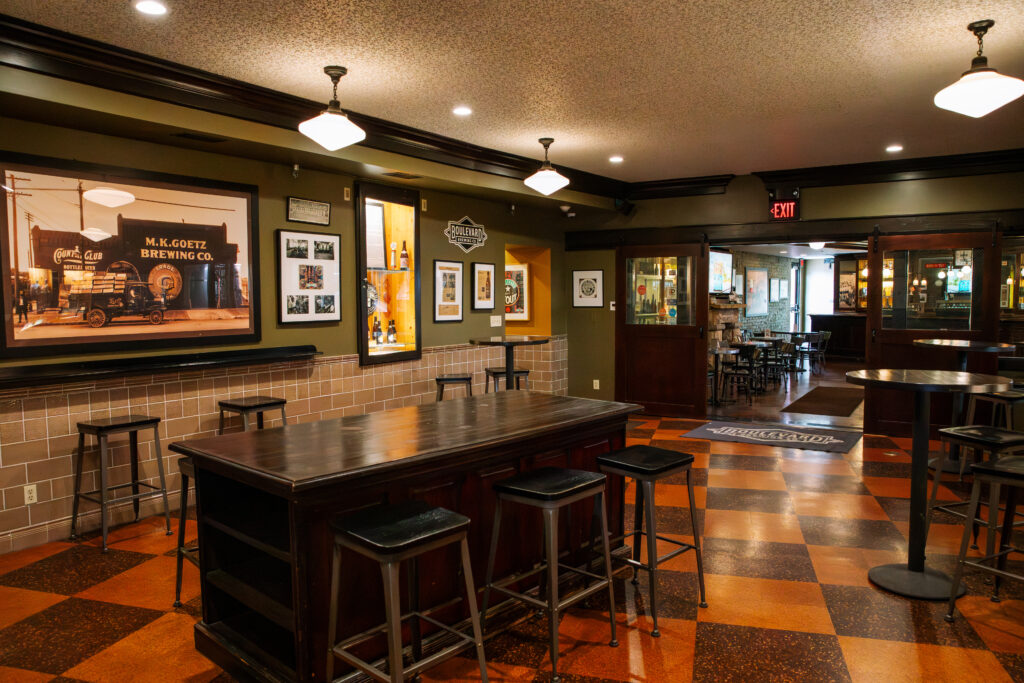 A cozy brewery taproom with dark wood tables and stools, vintage beer photos on the walls, checkered brown tile floor, and warm ceiling lights. An open doorway leads to more seating in the background.