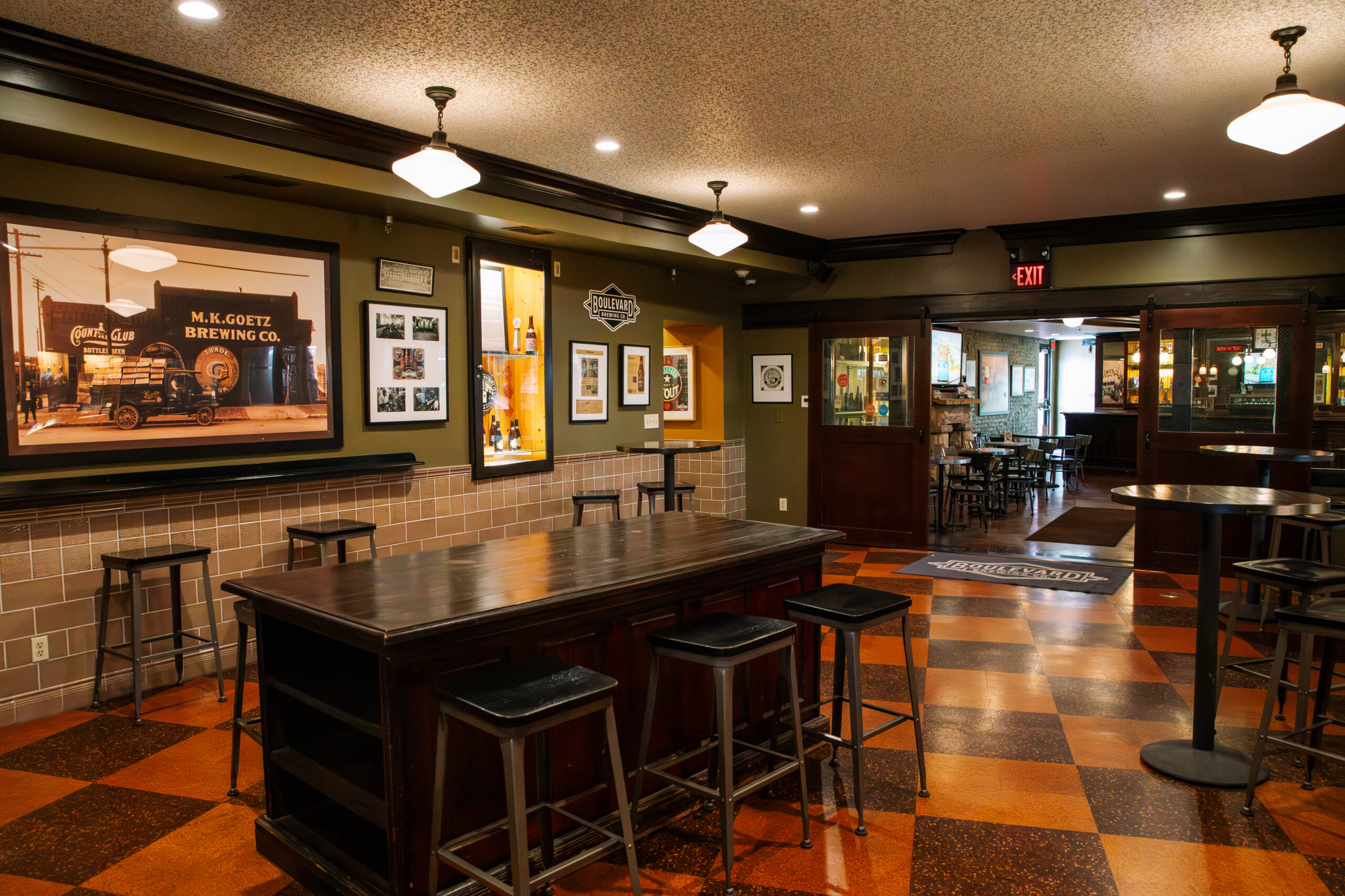 A cozy brewery taproom with dark wood tables and stools, vintage beer photos on the walls, checkered brown tile floor, and warm ceiling lights. An open doorway leads to more seating in the background.