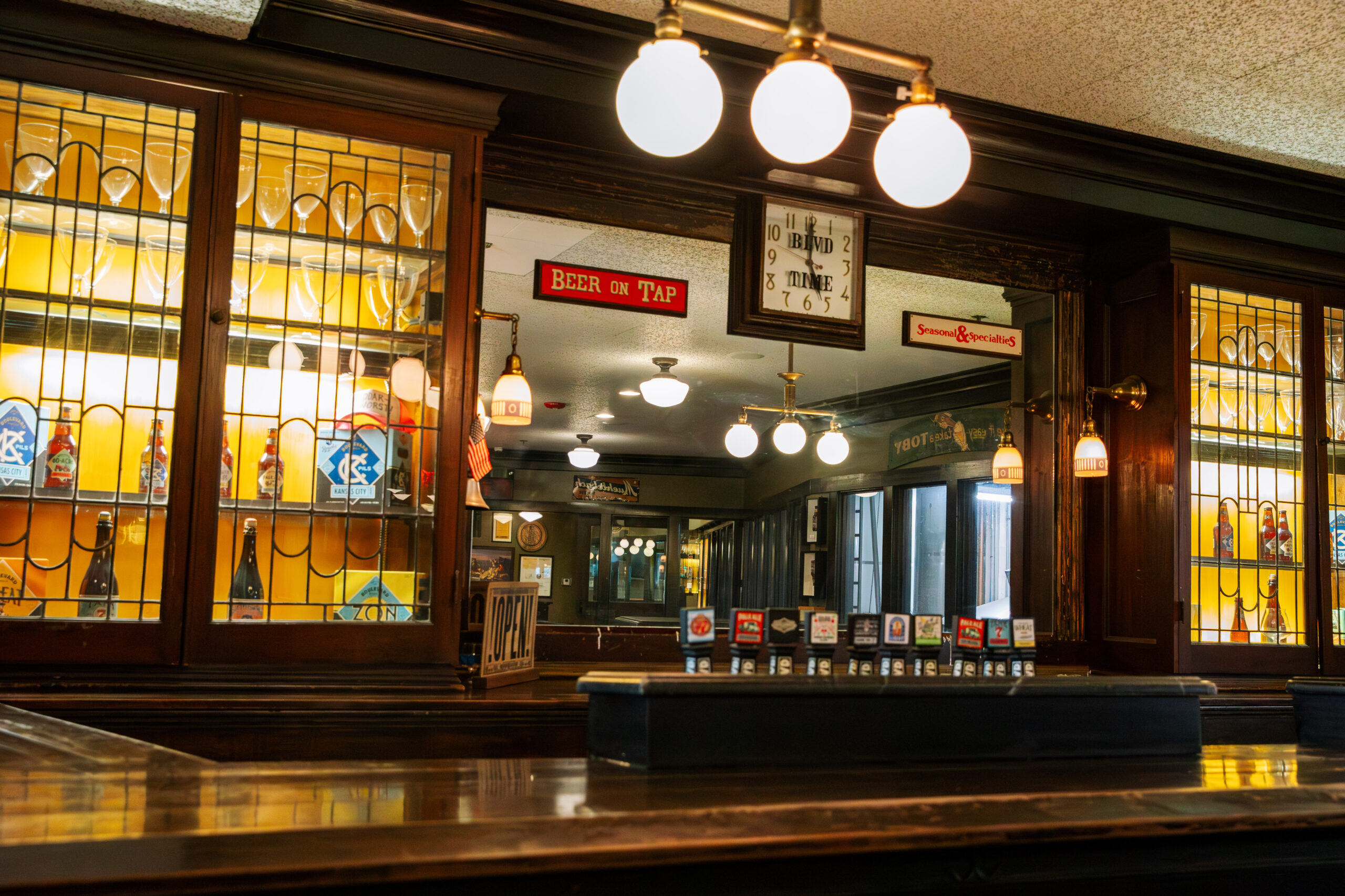 A vintage bar interior with wooden counters, stained glass cabinets displaying bottles, retro signs including "Beer on Tap," and round hanging lights reflected in a large mirror behind the bar.