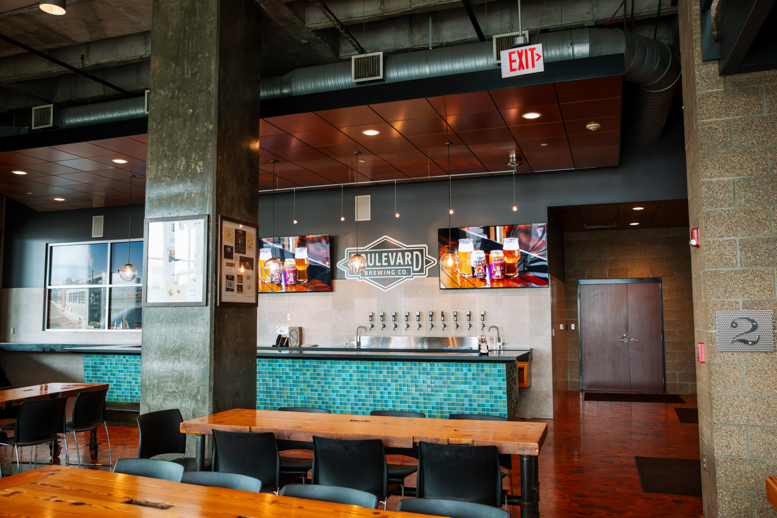 A modern brewery taproom with wooden tables, black chairs, and a bar featuring blue tiles and multiple beer taps. The wall displays "Boulevard Brewing Co." signage and large beer images.