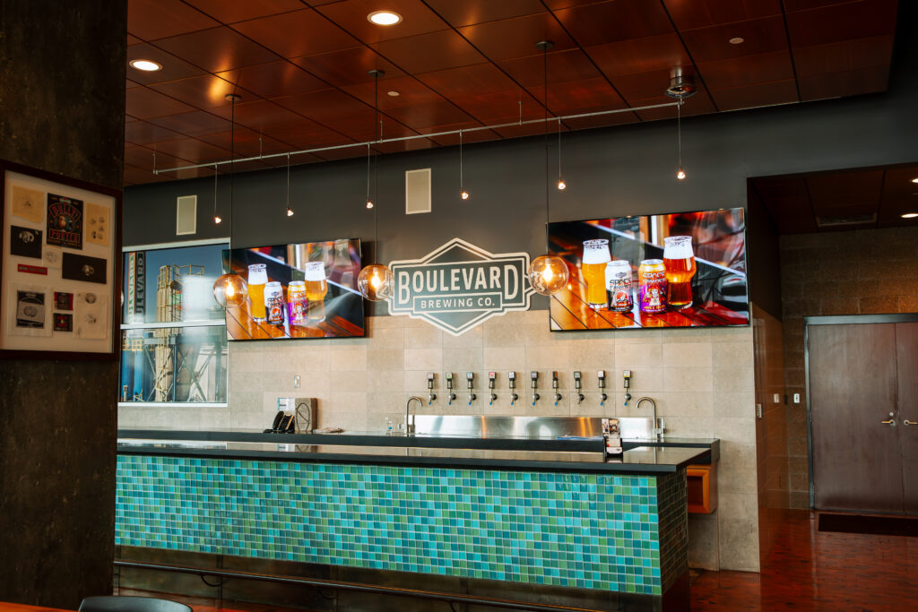 A modern brewery bar with a tiled counter, multiple beer taps, two TVs displaying images of beer, and a large "Boulevard Brewing Co." sign on the wall.