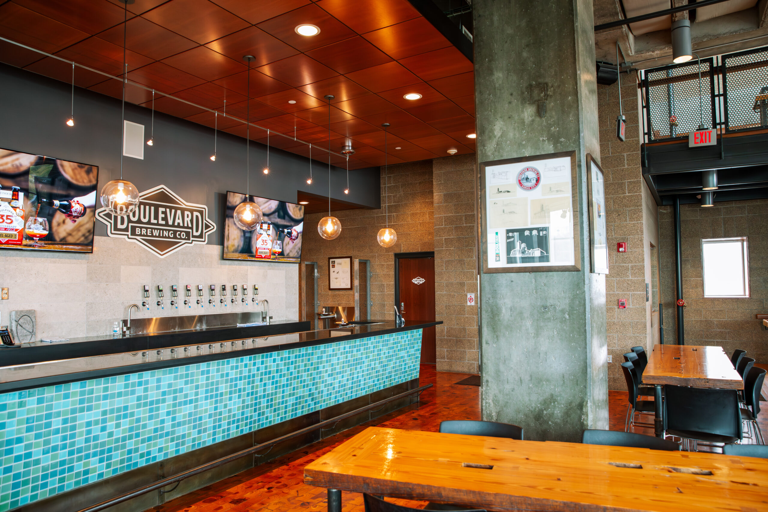 A modern brewery taproom with a tiled bar, Boulevard Brewing Co. sign, beer taps, hanging lights, wooden tables, TVs on the wall, and industrial-style decor.