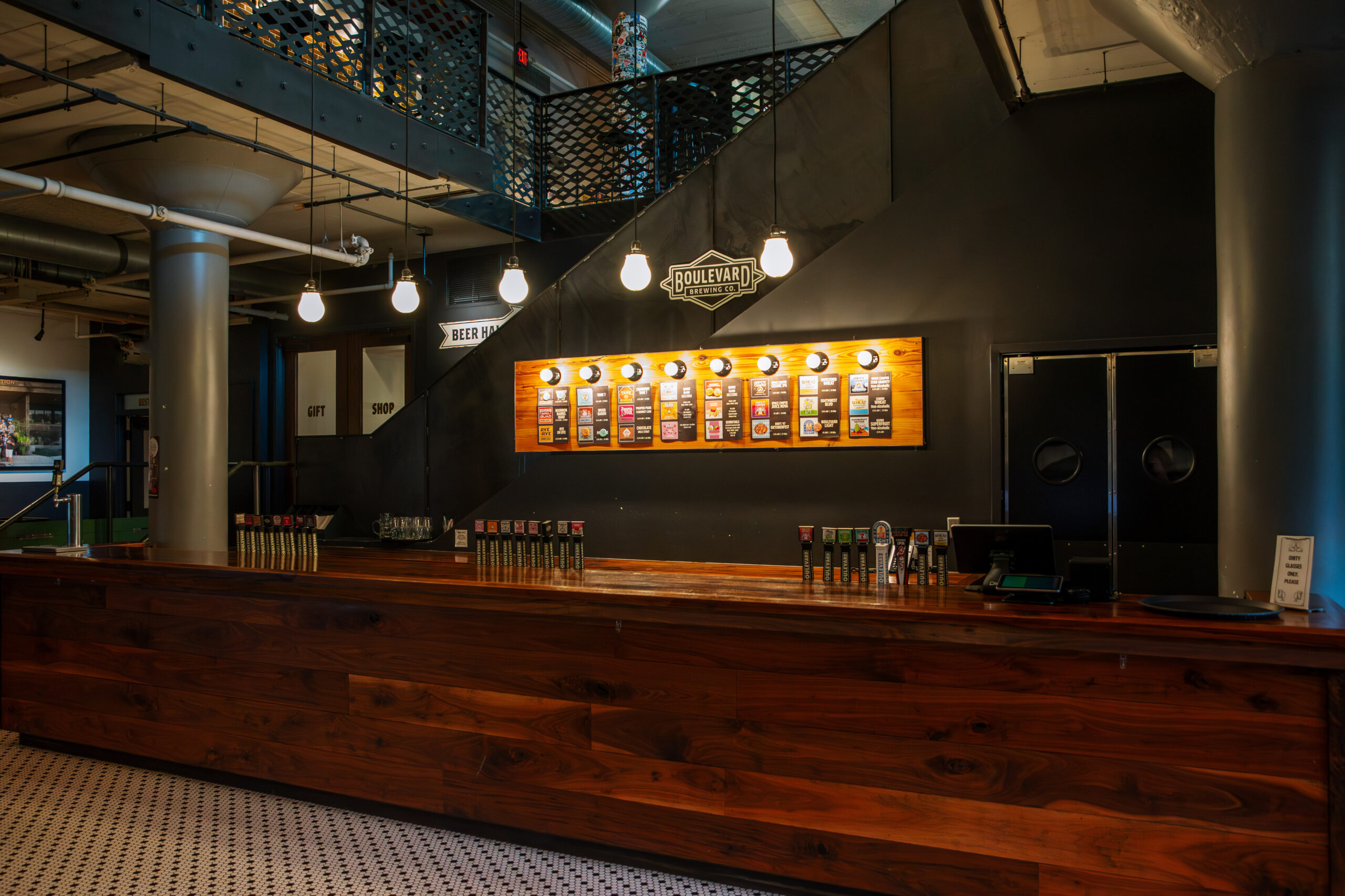 A modern bar with a long wooden counter, a wall-mounted beer menu, and hanging light bulbs. Stairs and industrial decor are visible, with black walls and white patterned floor tiles.