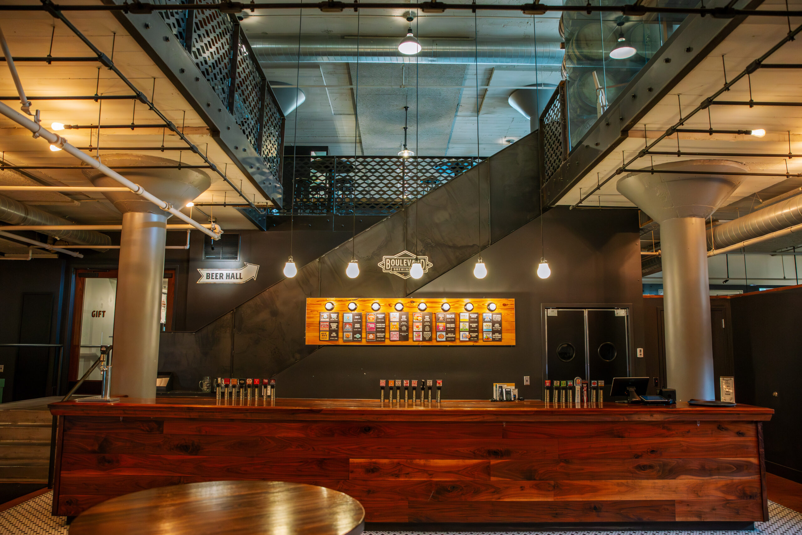 A modern brewery taproom with a long wooden bar, a beer menu on the wall, hanging lights, and an industrial-style staircase leading to an upper level. The space features exposed pipes and metal railings.