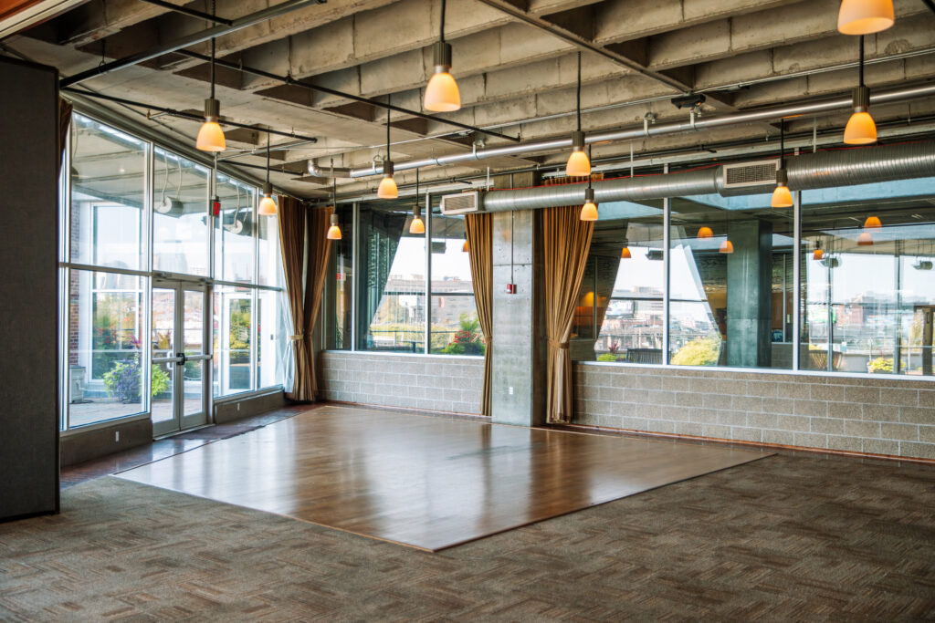 A spacious, modern room with large windows, exposed ceilings, hanging lights, carpeted floors, and a wooden dance floor area. Outside greenery and cityscape are visible through the glass walls.