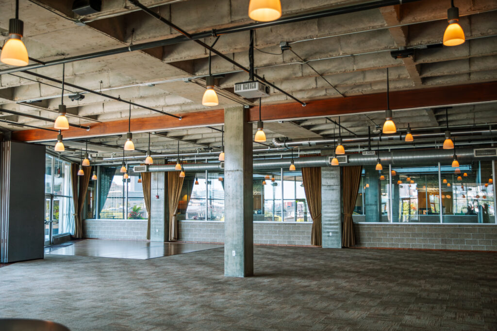 Spacious, modern industrial room with carpeted floors, exposed concrete ceiling, hanging pendant lights, large windows, and brown curtains, offering an open and versatile event or meeting space.