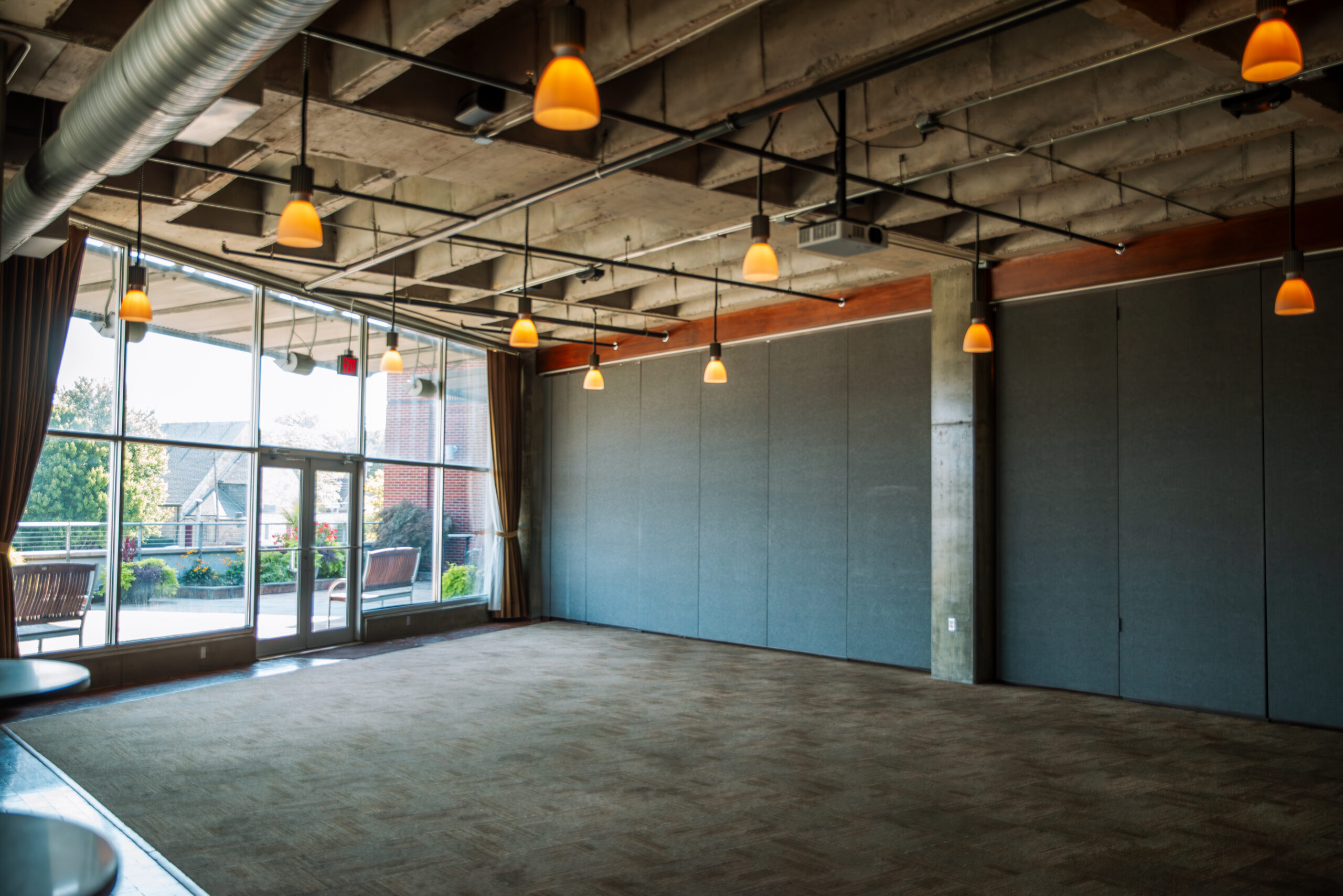 Spacious, empty room with carpeted floors, exposed ceiling with hanging lights, large windows letting in natural light, and a view of trees and a patio outside.