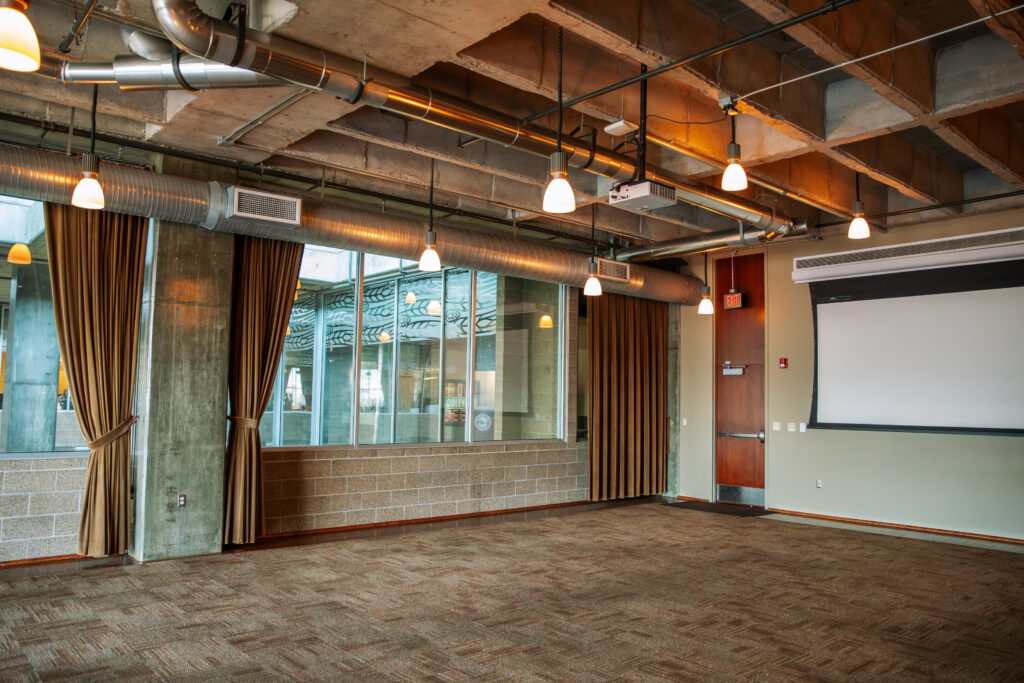 A modern, empty conference room with carpeted floors, large windows with brown curtains, exposed ceiling pipes, hanging lights, a wall-mounted projector screen, and a glass wall overlooking an interior courtyard.