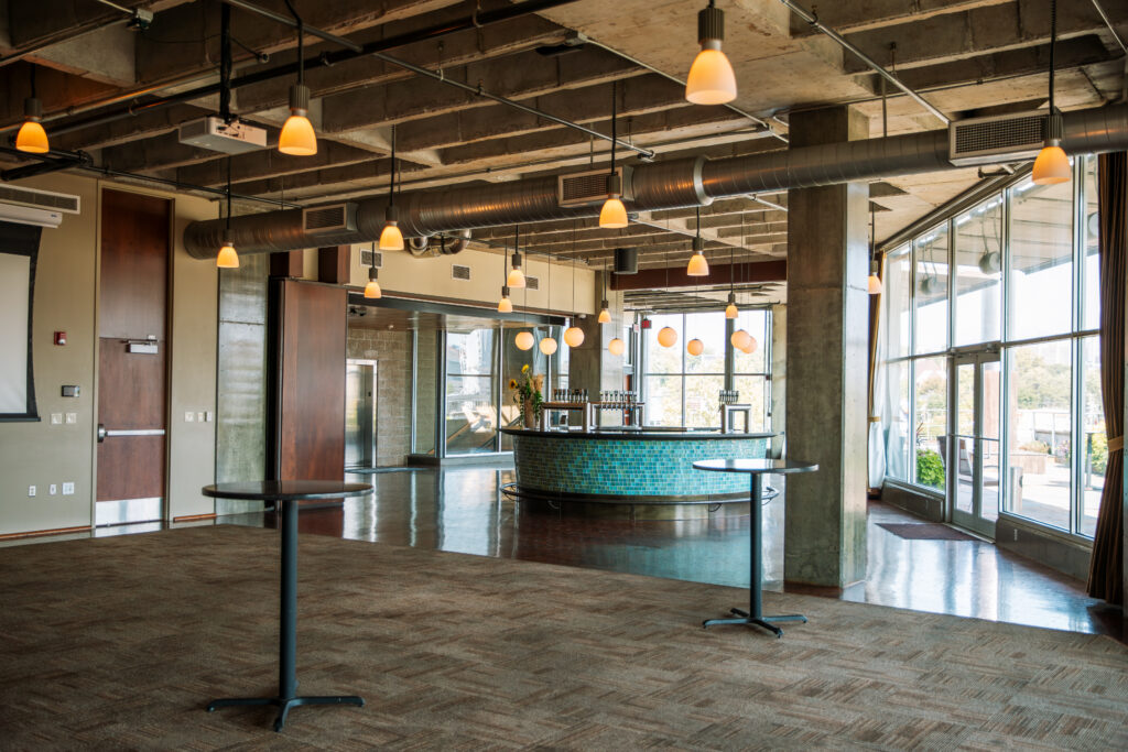 A spacious, modern event venue with exposed ceiling, pendant lights, large windows, a tiled bar with barstools, and empty high-top tables on a carpeted floor. Natural light fills the open area.