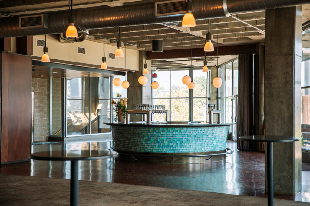 A modern, empty bar area with a curved counter covered in blue tiles, hanging pendant lights, and large windows letting in natural light. The space has exposed ductwork and a polished floor.