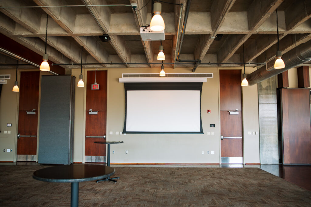An empty conference room with a blank projector screen, several round tables, pendant lights hanging from the ceiling, and exposed concrete beams and ducts.