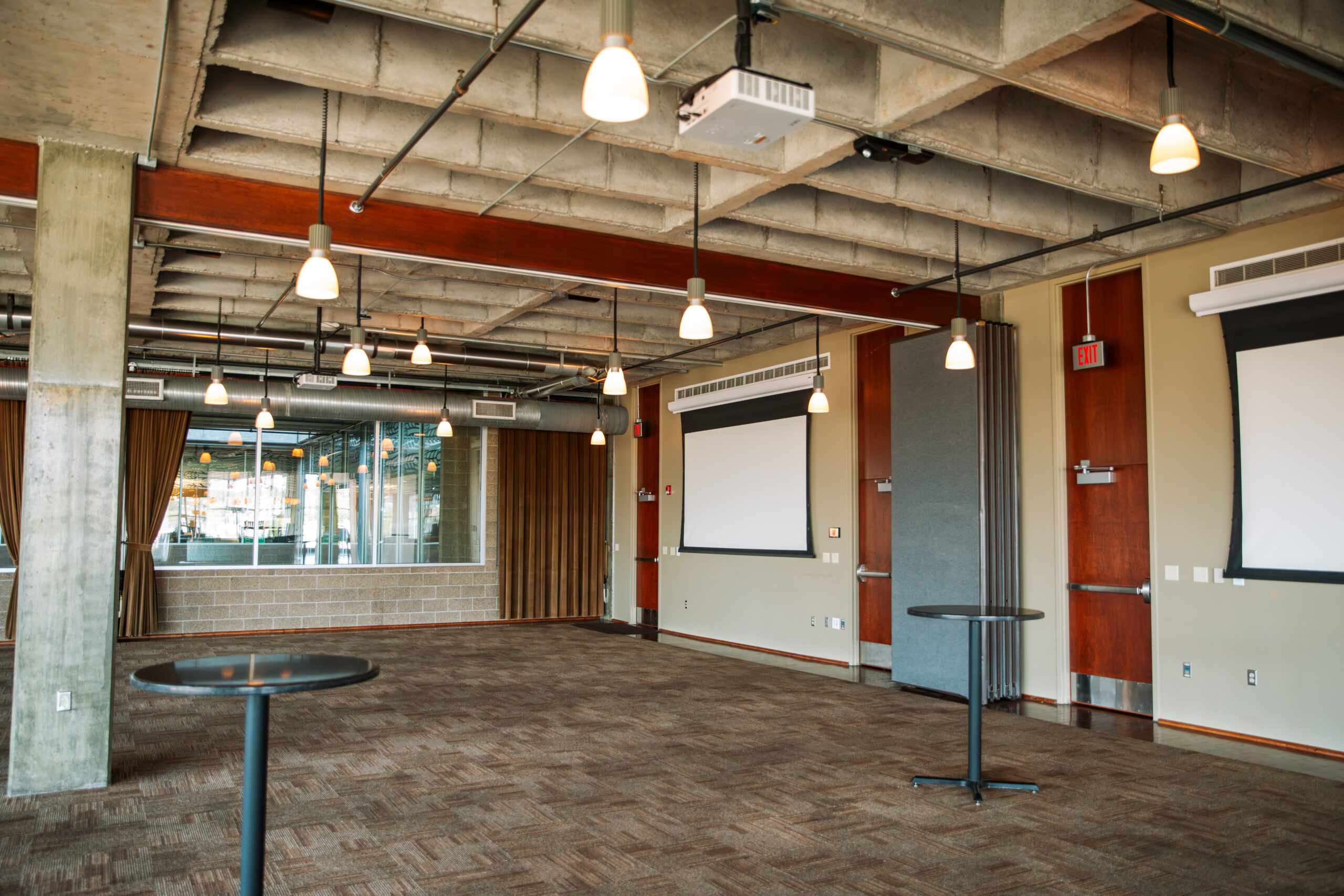 Spacious, modern conference room with exposed concrete ceiling, hanging lights, large windows, two retractable projector screens, and a few tall round tables on a carpeted floor.