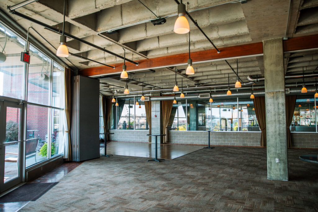 Spacious modern room with large windows, exposed concrete ceiling, hanging lights, carpeted floor, and mirrored walls. The room appears empty and well-lit with natural light.