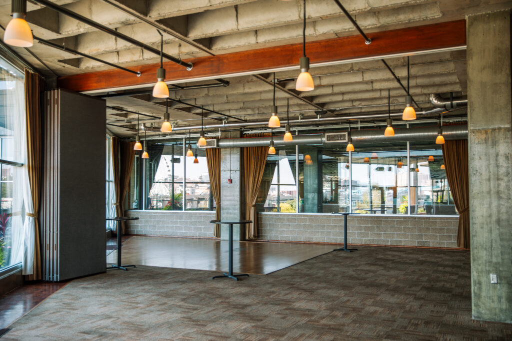 Spacious, modern event room with large windows, exposed ceilings, hanging lights, carpeted and wooden flooring, gold curtains, and cocktail tables. Natural light fills the area through the glass walls.