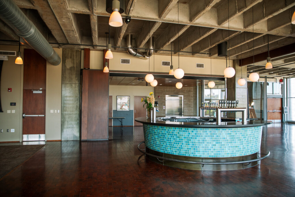 A modern, spacious room with an industrial ceiling, hanging pendant lights, a circular bar covered in blue tiles, and large windows letting in natural light. The room has wooden floors and metal air ducts.