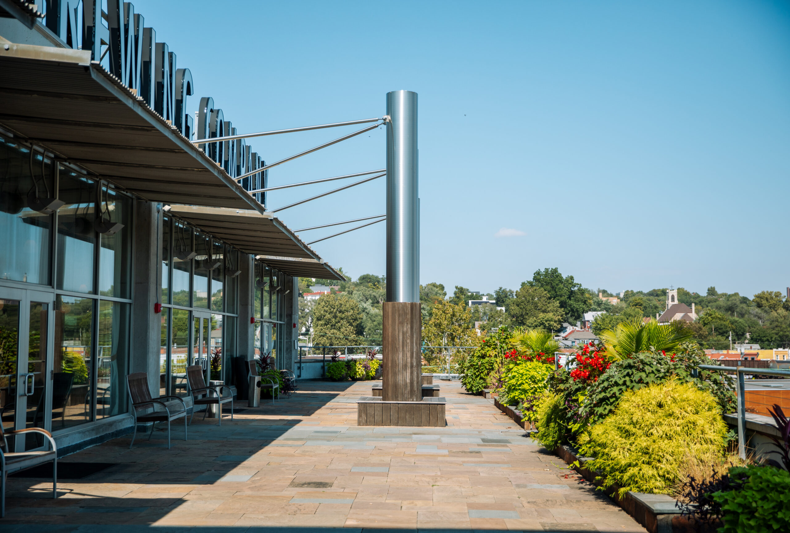 A sunlit outdoor terrace with benches, potted plants, and large modern metal columns next to a building with glass walls. Trees and buildings are visible in the background under a clear blue sky.