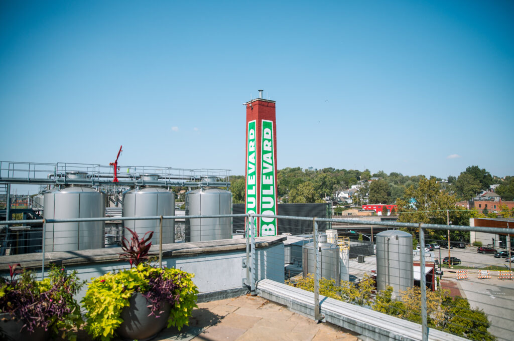 A rooftop view features potted plants in the foreground, large metal brewery tanks, and a tall red sign reading "Boulevard" in green and white, with buildings and trees visible in the background under a clear blue sky.