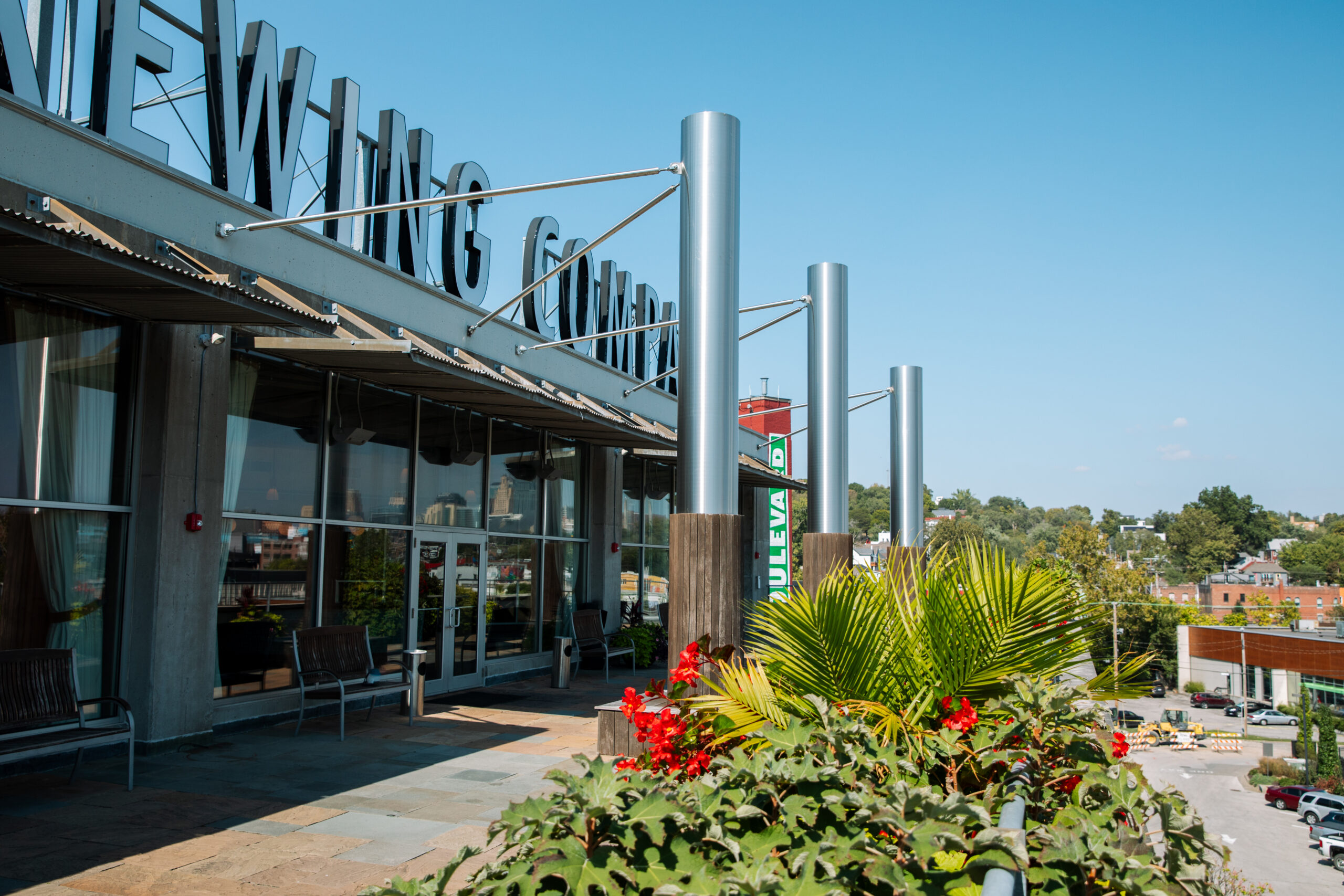 A modern building with large glass windows and a metal overhang is shown on a sunny day. Green plants with red flowers are in the foreground; a parking lot and trees are visible in the background.