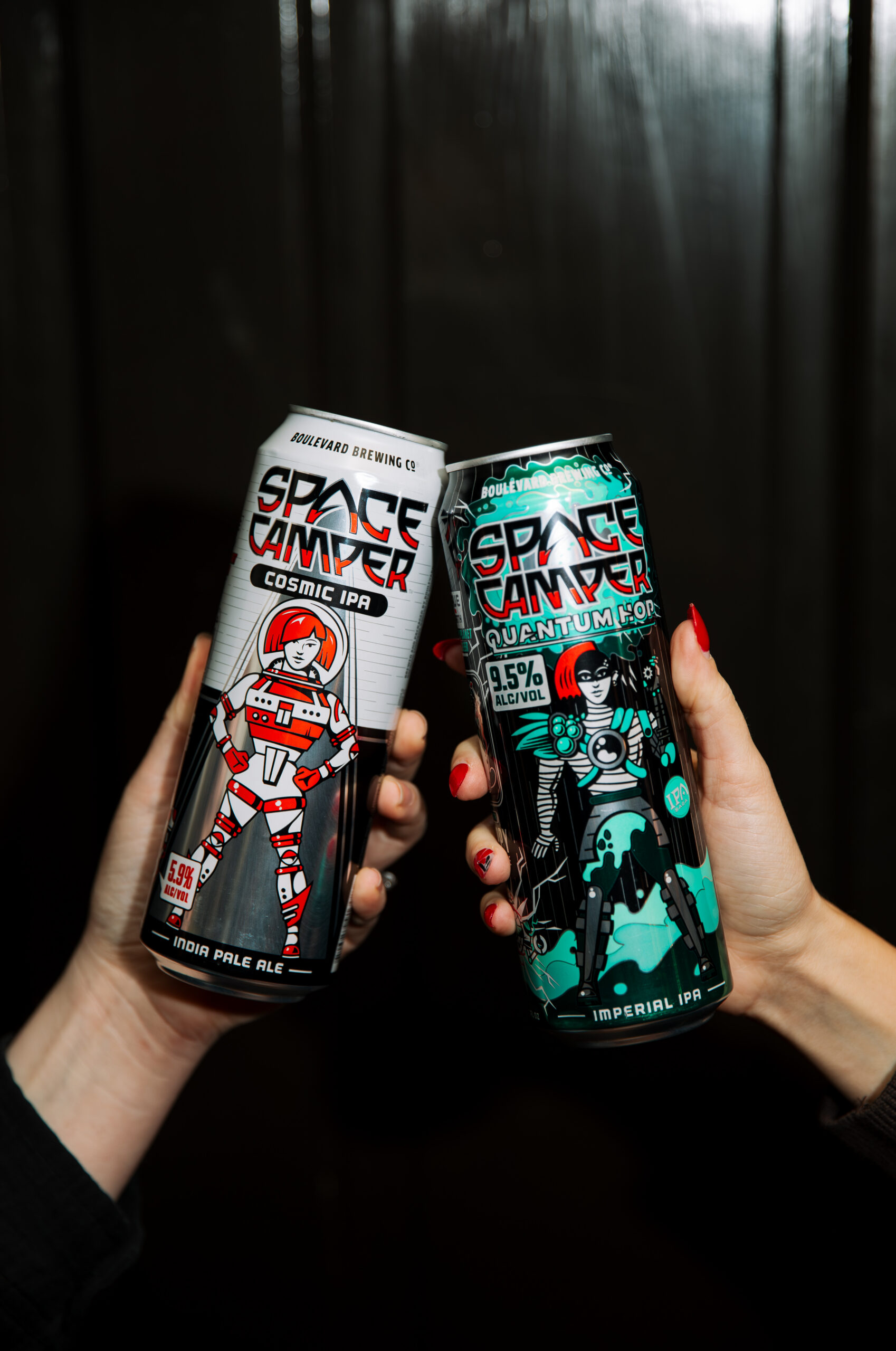 Two hands hold up cans of Space Camper IPA beer—one labeled “Cosmic IPA” with a red and white astronaut, and the other labeled “Quantum Fog” featuring a teal astronaut—set against a dark background. The distinctive Space Camper design boldly highlights its cosmic inspirations.