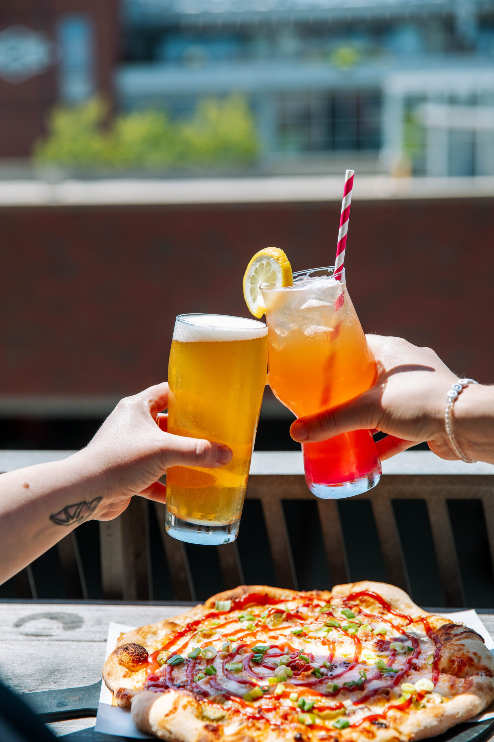Two people clink a glass of Boulevard Brewing Co. Beer Hall beer and a colorful cocktail with a lemon slice and striped straw over a table with freshly baked pizza, outdoors at the Tours and Rec Center in Kansas City on a sunny day.