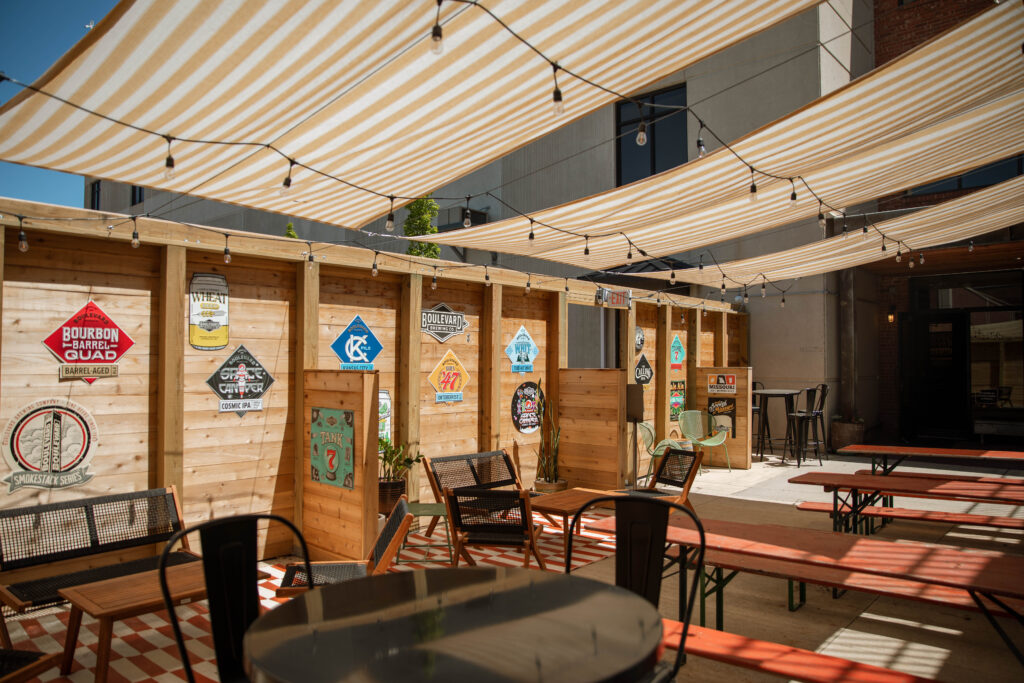 Outdoor patio with striped shade canopies, wooden fence decorated with colorful vintage signs, and red picnic tables with benches. The area has chairs and tables, creating a casual, inviting atmosphere.