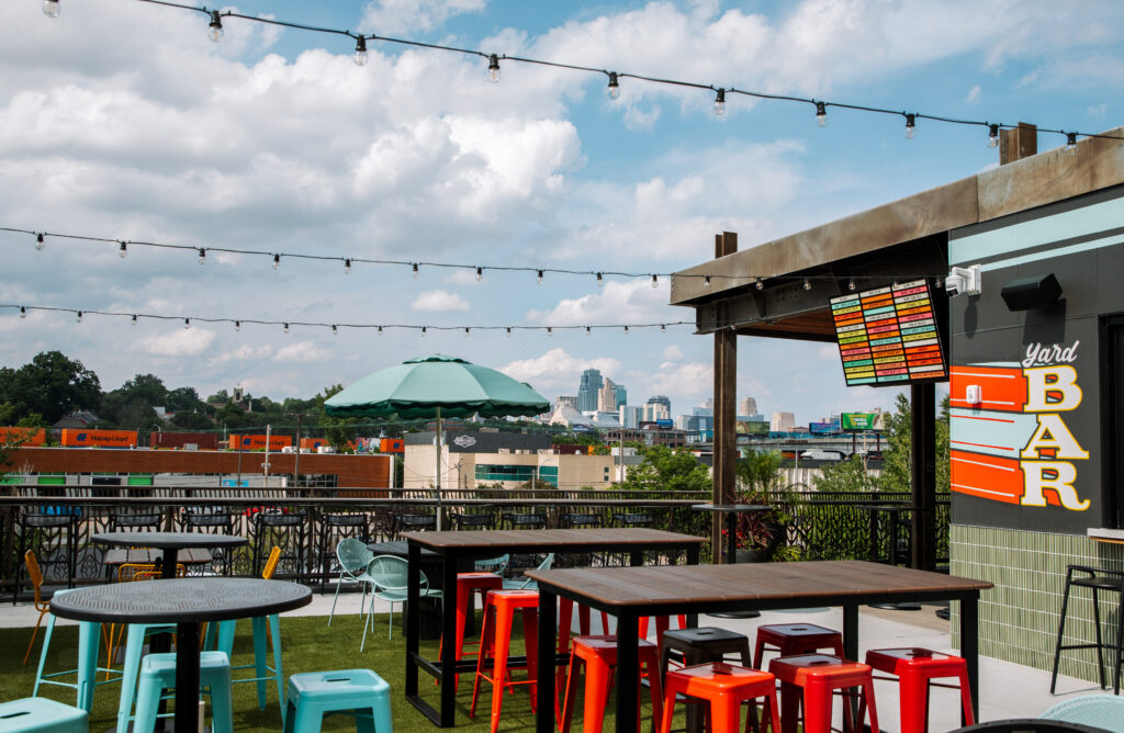 Rooftop patio at the Boulevard Brewing Co. Beer Hall with high-top tables, red stools, a green umbrella, and a digital menu board overlooking a distant Kansas City skyline.