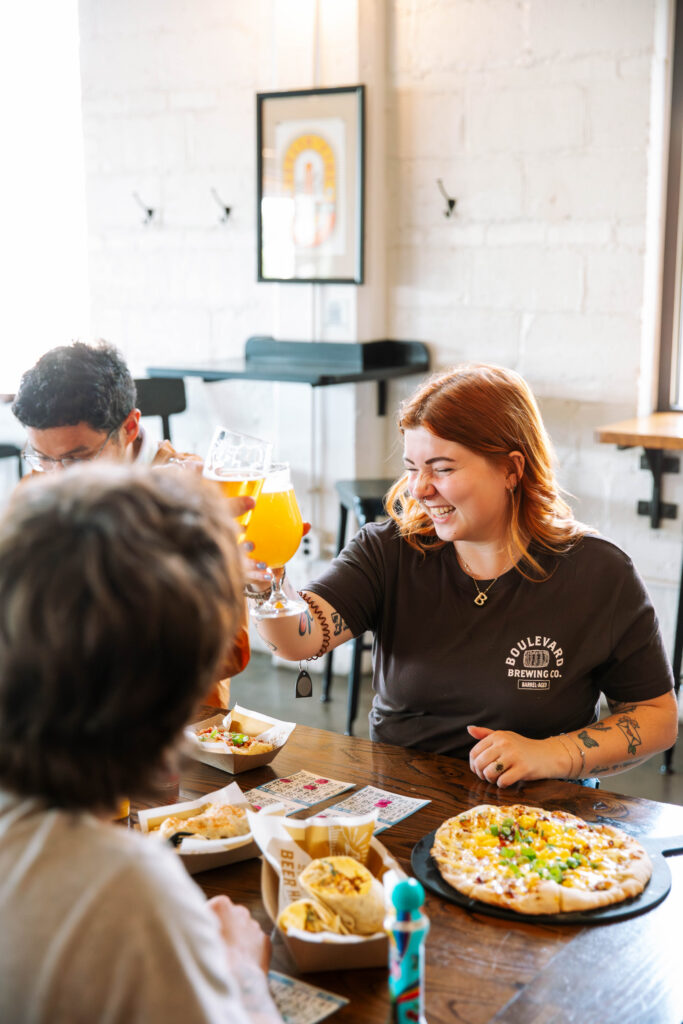 Three friends laugh while clinking glasses of Boulevard Brewing Co. beer at a restaurant table with pizza and appetizers.