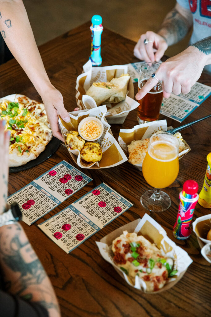 People eating pizza and snacks while playing bingo at a wooden table with glasses of beer at the Boulevard Brewing Co. Beer Hall.