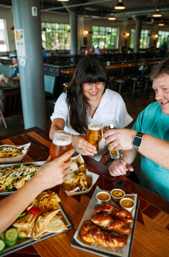 Three people clinking beer glasses together over a table spread with nachos, pretzels, and dips at the Boulevard Brewing Co. Beer Hall.